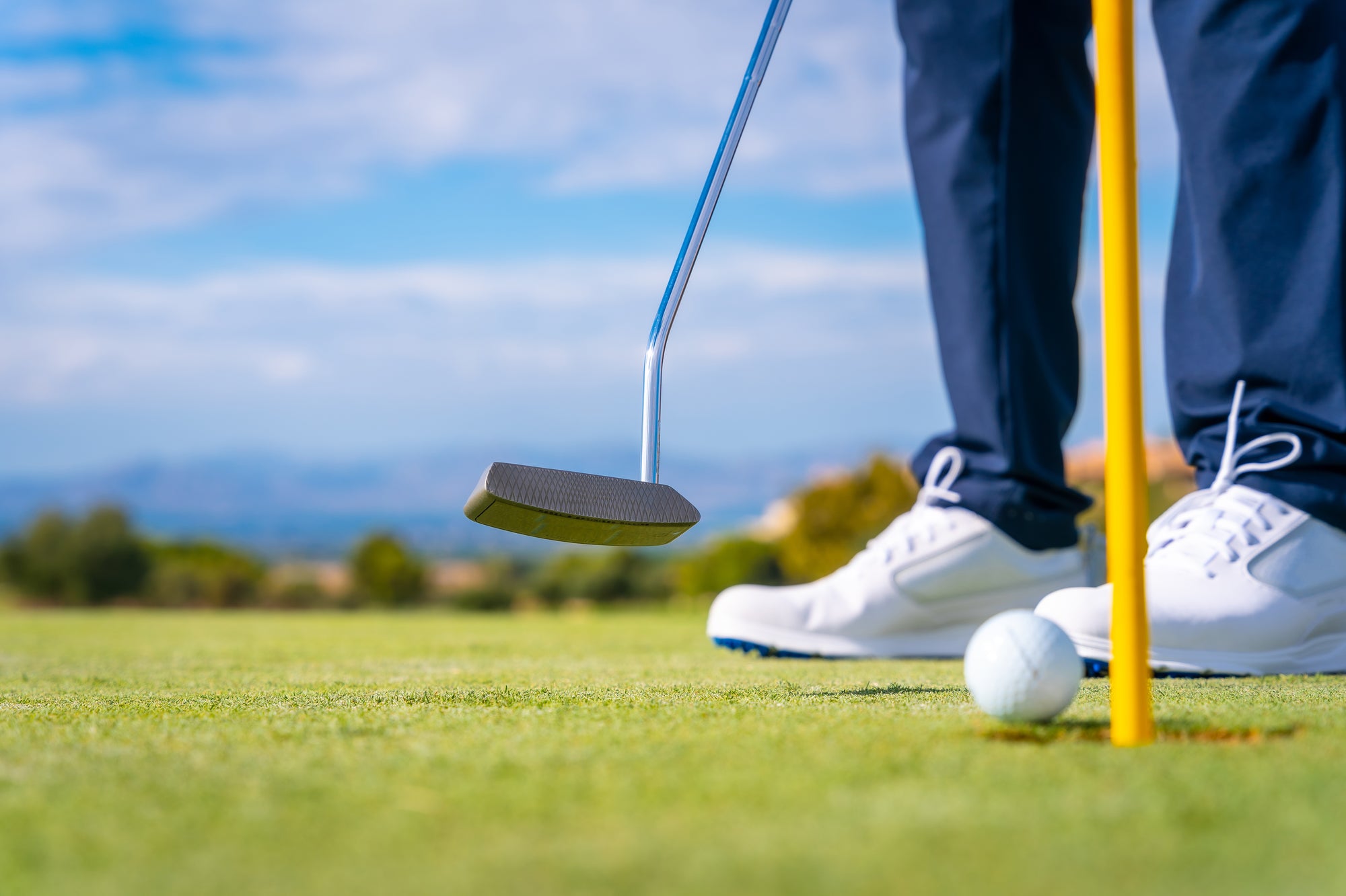A close-up action shot of a golfer's lower legs and feet wearing white golf shoes, holding a silver putter above a golf ball on a yellow flagstick, set against a blurred background of a golf course and blue sky.